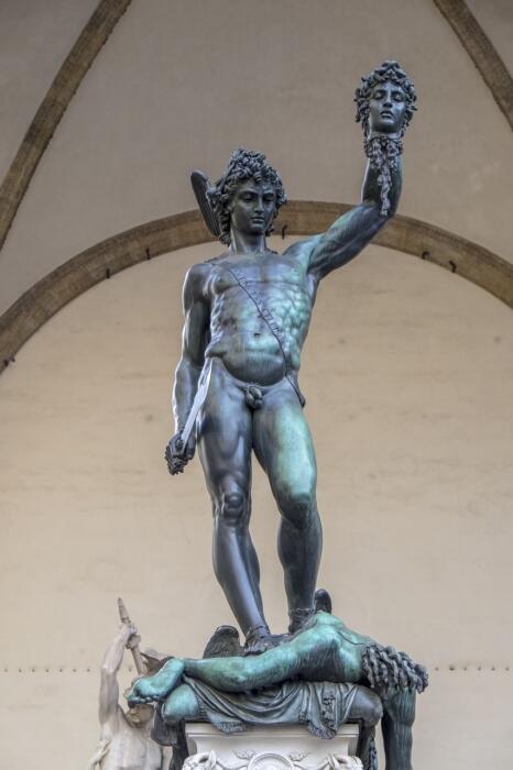 A bronze statue of a muscular nude man standing triumphantly on a pedestal, holding a severed head aloft in his right hand, conveying victory and strength.