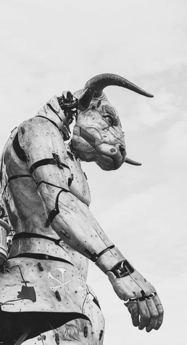 Large mechanical bull with humanoid features, constructed from metal plates and bolts, stands outdoors against a cloudy sky in black and white.